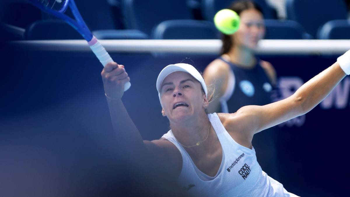 Tenisistka w białej koszulce i czapce z daszkiem wykonuje zamach rakietą, koncentrując się na uderzeniu piłki tenisowej. W tle widoczna inna zawodniczka.