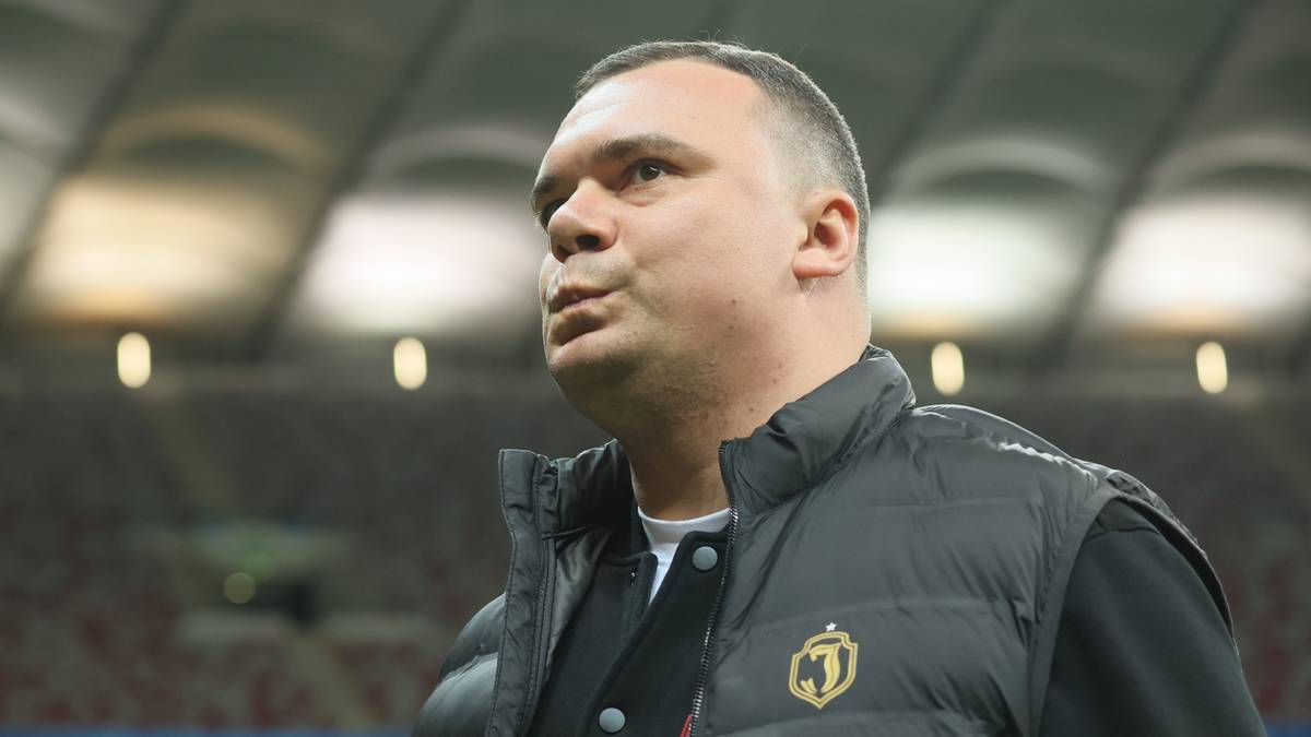 Mężczyzna w kamizelce z herbem klubu piłkarskiego na tle stadionu.