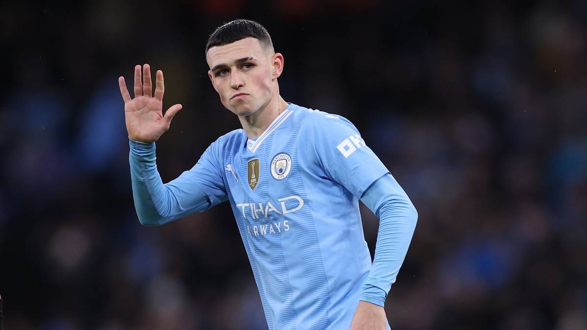 Piłkarz Manchesteru City, Phil Foden, w niebieskiej koszulce, unosi prawą dłoń otwartą w geście pozdrowienia.