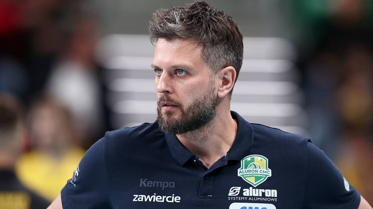 Zbliżenie na twarz Michała Winiarskiego, siatkarza i trenera, noszącego granatową koszulkę z logo Aluron i napisem "Kempa Zawiercie". Włosy ciemne, krótko ścięte, z lekko rozwichrzoną grzywką. Broda krótka, kilkudniowa. Patrzy w lewo, z lekkim zamyśleniem na twarzy. Tło jest rozmyte, sugerujące halę sportową z widocznymi sylwetkami innych osób.