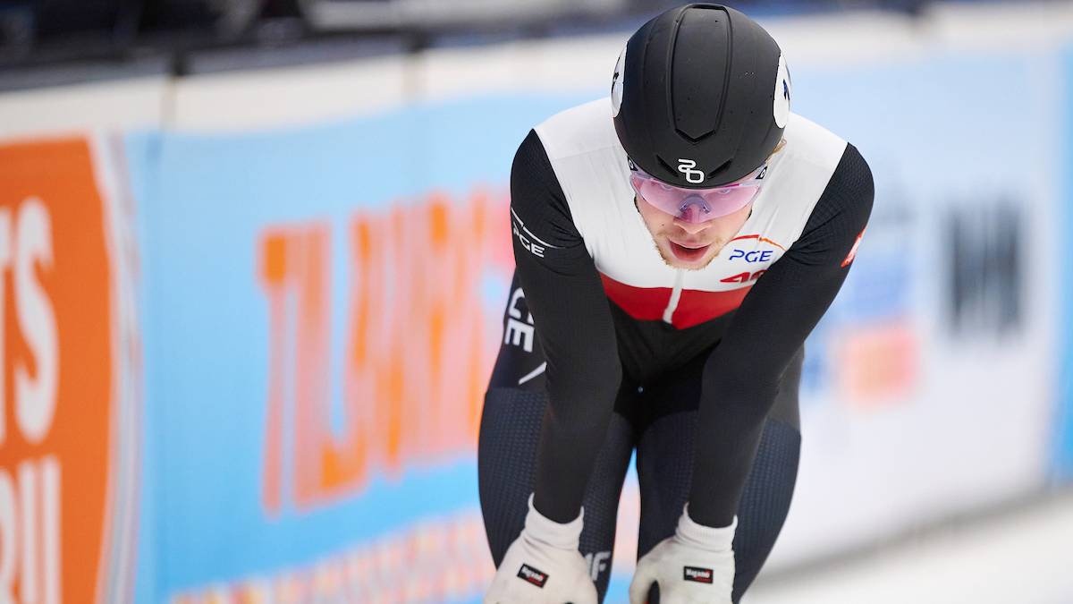 Zawodnik short tracku w kasku i kombinezonie pochyla się na lodowisku, opierając jedną rękę na kolanie.