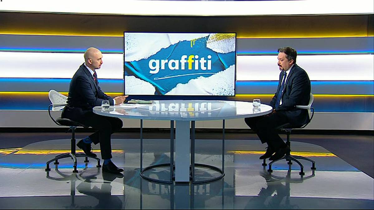 Dw&oacute;ch mężczyzn w garniturach siedzi przy okrągłym stole w studiu telewizyjnym, naprzeciwko siebie. Za nimi znajduje się duży ekran z napisem "graffiti".