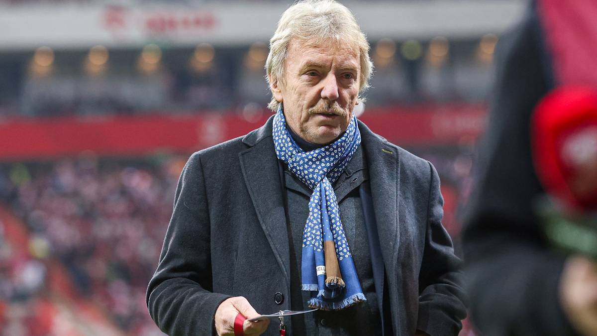 Mężczyzna w średnim wieku z siwymi włosami, ubrany w ciemny płaszcz i niebieski szalik w białe kropki, trzyma w dłoni nożyczki.