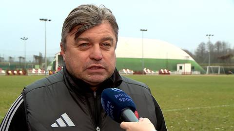 Roman Kosecki udziela wywiadu na tle boiska piłkarskiego. Ma na sobie ciemną kamizelkę z białymi paskami, przed nim trzymany jest mikrofon.