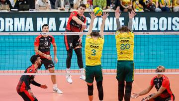 Siatkarze w akcji podczas meczu, dw&oacute;ch graczy w ż&oacute;łtych strojach pr&oacute;buje zablokować atak przeciwnika w czerwono-czarnym stroju.