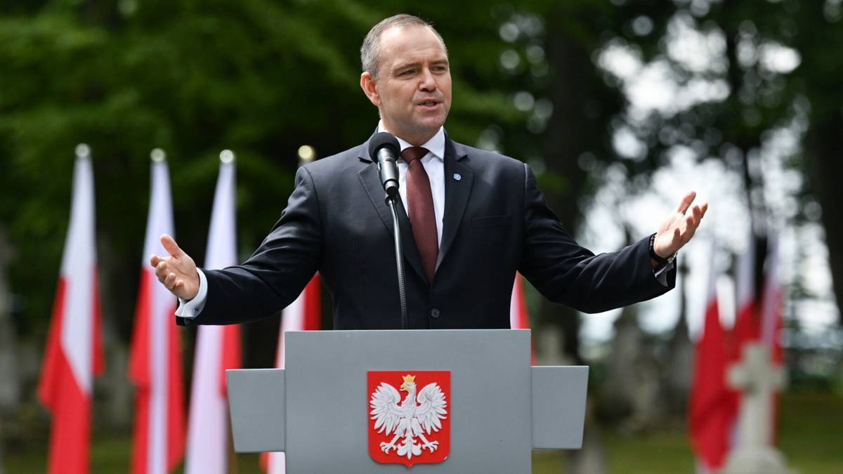 Mężczyzna w garniturze przemawia za mównicą z godłem Polski.