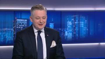 Mężczyzna w garniturze i krawacie siedzi na tle niebieskiego ekranu z widokiem nocnego miasta.