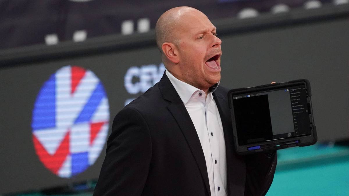 Mężczyzna w garniturze i koszuli, trzymający tablet, krzyczy na boisku siatkarskim.