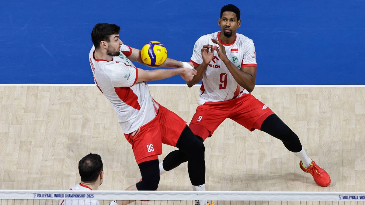 Dwóch zawodników w białych koszulkach z czerwonymi elementami próbuje zablokować atak przeciwnika podczas meczu siatkówki.