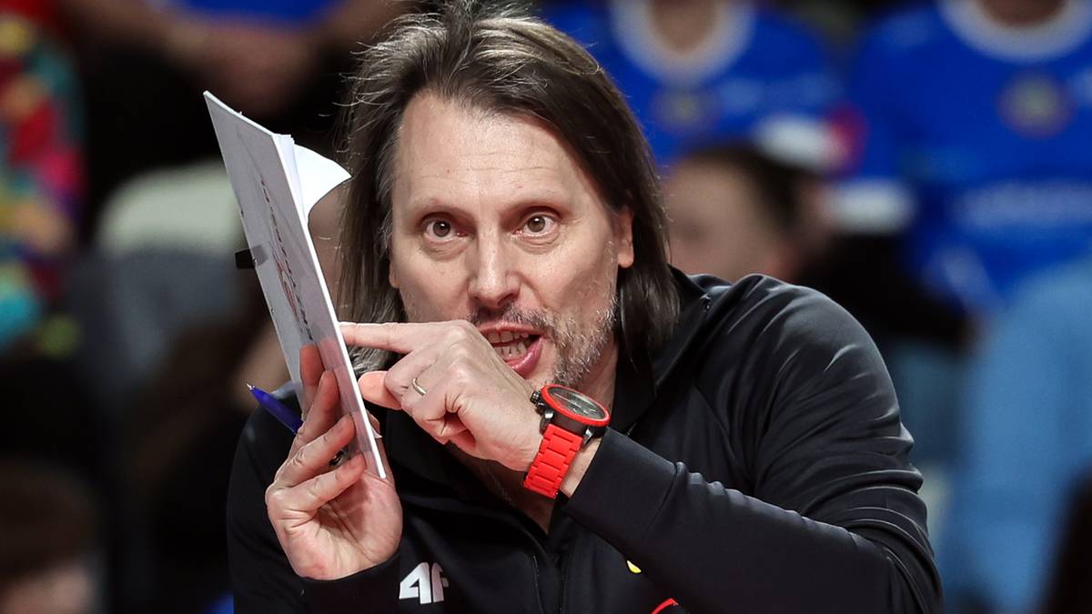 Trener siatkówki z kartką w ręku, wskazujący palcem, na tle publiczności.