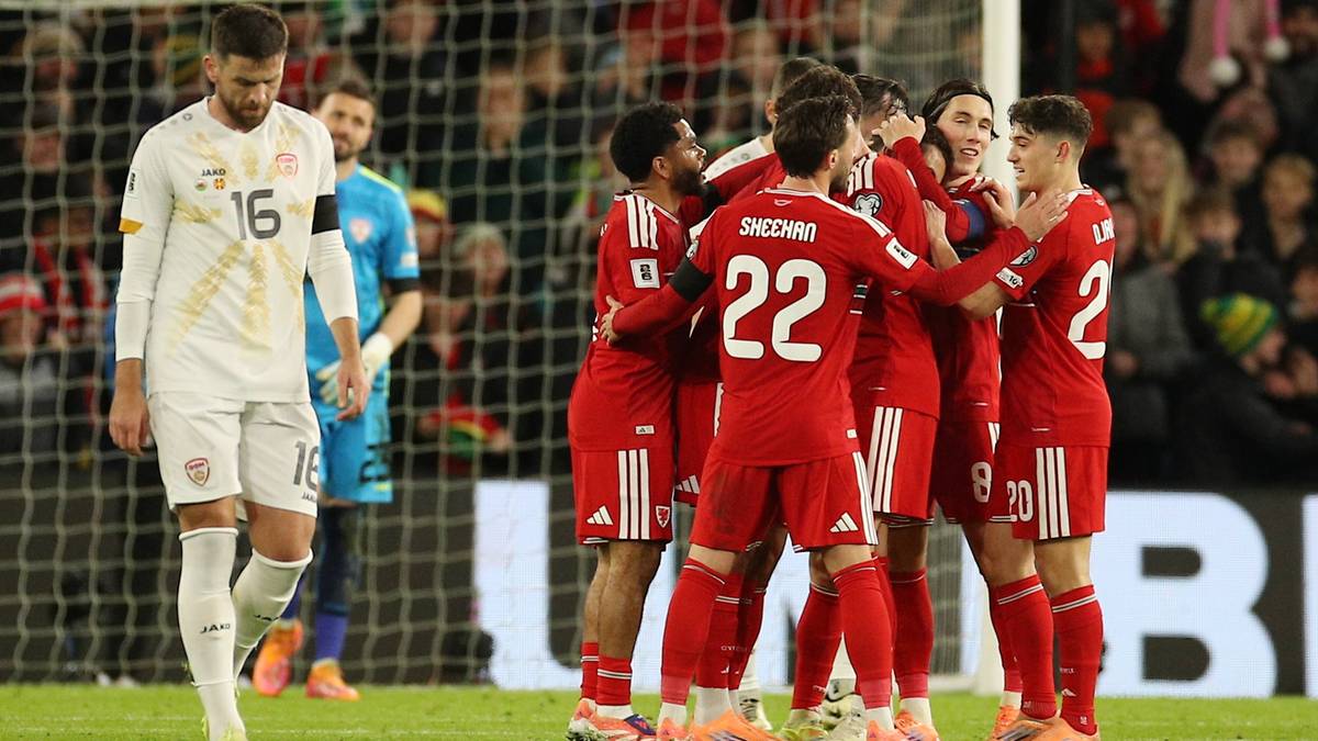 Piłkarze w czerwonych strojach cieszą się ze zdobytego gola, jeden z zawodników w białym stroju odchodzi zrezygnowany.