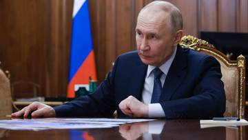 Władimir Putin siedzi przy stole w gabinecie, za nim flaga Rosji.