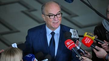 Mężczyzna w niebieskim garniturze i okularach stoi otoczony przez mikrofony.