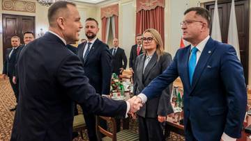 Dw&oacute;ch mężczyzn w garniturach, jeden z nich minister, ściska sobie dłonie, podczas gdy inni obserwują.