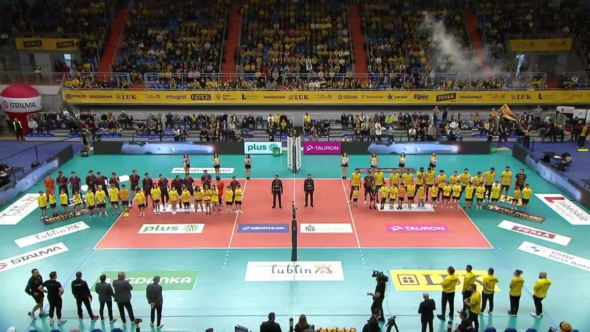 Boisko do siatk&oacute;wki z dwiema drużynami ustawionymi po przeciwnych stronach, w tle widownia wypełniona kibicami.