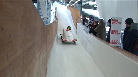 Dwie kobiety w kasku, jadące w białym bobsleju, zjeżdżają po lodowym torze.