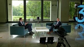Dw&oacute;ch mężczyzn w garniturach siedzi naprzeciwko siebie na kanapach w studiu telewizyjnym. Za nimi znajdują się duże okna z widokiem na zieleń.
