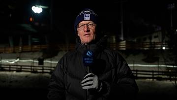 Mężczyzna w zimowej czapce i okularach z mikrofonem w ręku, stojący na zewnątrz w nocy.