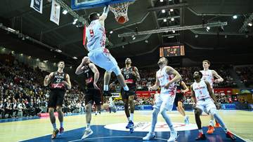 Gracz w białej koszulce z numerem 9 wykonuje wsad do kosza podczas meczu koszyk&oacute;wki. Otaczają go inni zawodnicy w czerwonych i białych strojach.