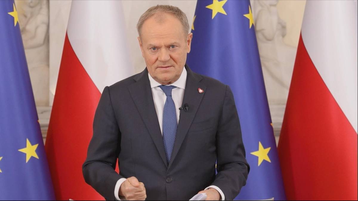 Premier Donald Tusk stoi przed flagami Polski i Unii Europejskiej, wygłaszając przem&oacute;wienie.