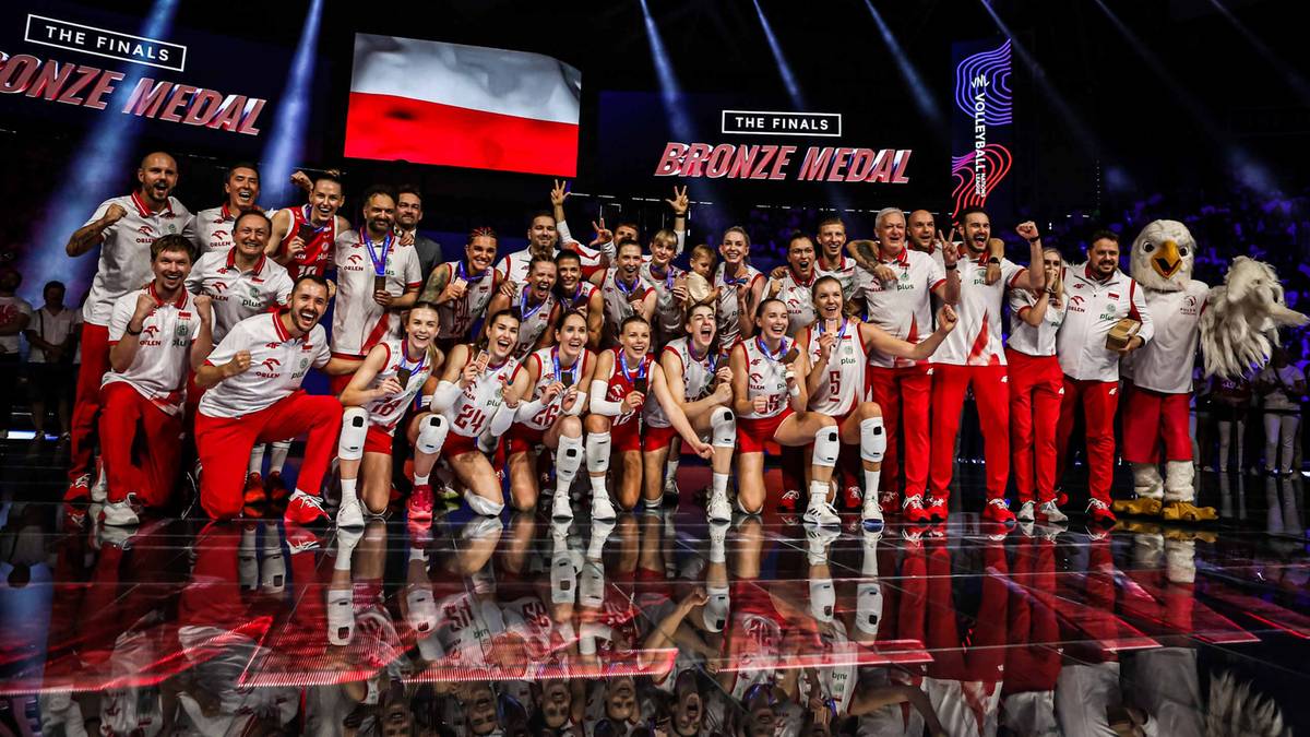 Drużyna siatkarek w strojach reprezentacyjnych pozuje do zdjęcia na podium, trzymając w rękach brązowy medal i polską flagę.