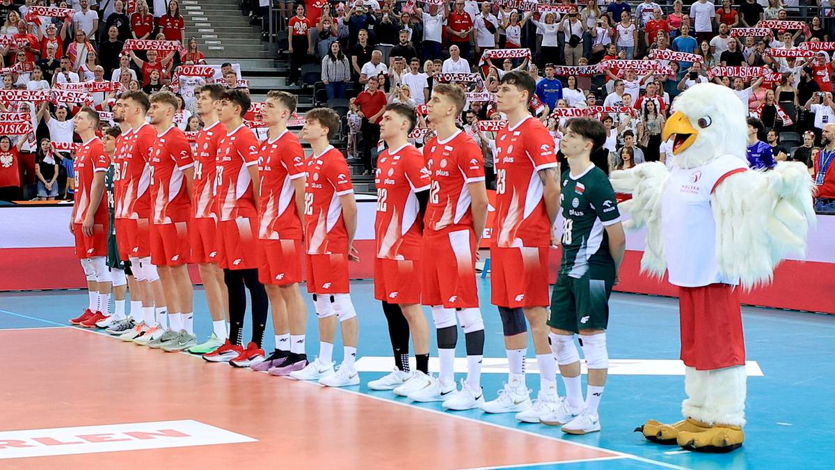 Drużyna siatkarzy Polski w czerwonych strojach stoi w rzędzie na boisku, w tle publiczność, obok postać w stroju maskotki orła.