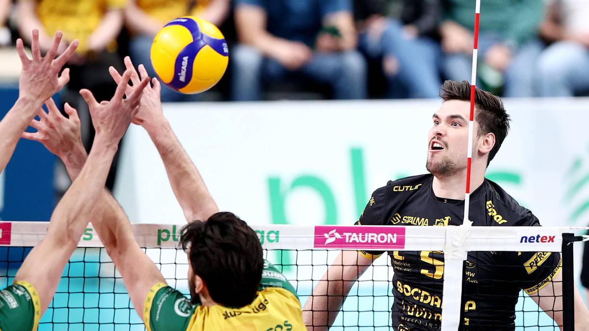Siatkarze walczą o piłkę nad siatką, jeden pr&oacute;buje ją odbić, drugi blokuje.