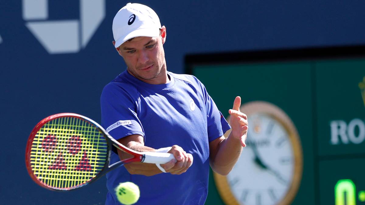 Mężczyzna w niebieskiej koszulce i białej czapce gra w tenisa, uderzając piłkę rakietą.