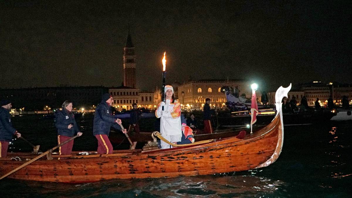 Ludzie wiosłują w gondoli z pochodnią na wodzie, z budynkami i wieżą w tle nocą.