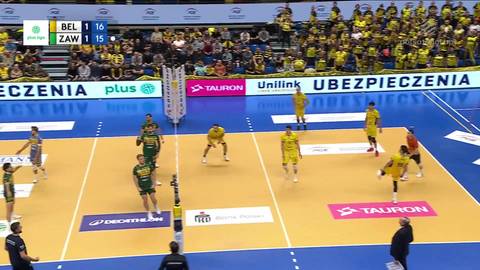 Gracze w żółtych i zielonych strojach grają w siatkówkę na boisku. Widoczni są kibice na trybunach.