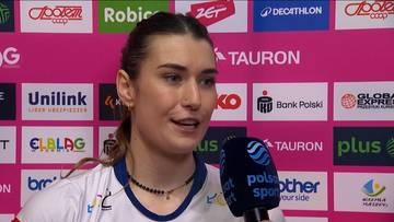 Portret młodej kobiety, siatkarki, z mikrofonem przed twarzą. Na jej szyi wisi naszyjnik z koralik&oacute;w. Tło stanowi ściana z logo sponsor&oacute;w.