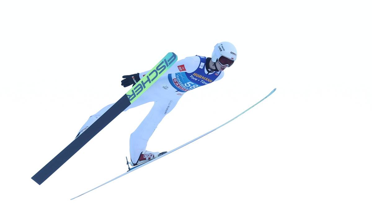 Skoczek narciarski w białym kombinezonie wykonuje skok, z nartami skierowanymi do przodu, na tle białego, jednolitego tła.