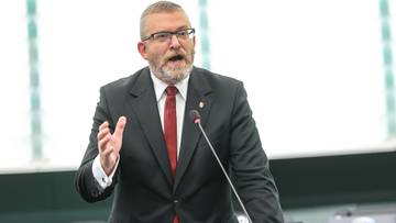 Mężczyzna w garniturze i okulary przemawia z m&oacute;wnicy, wykonując gest ręką.