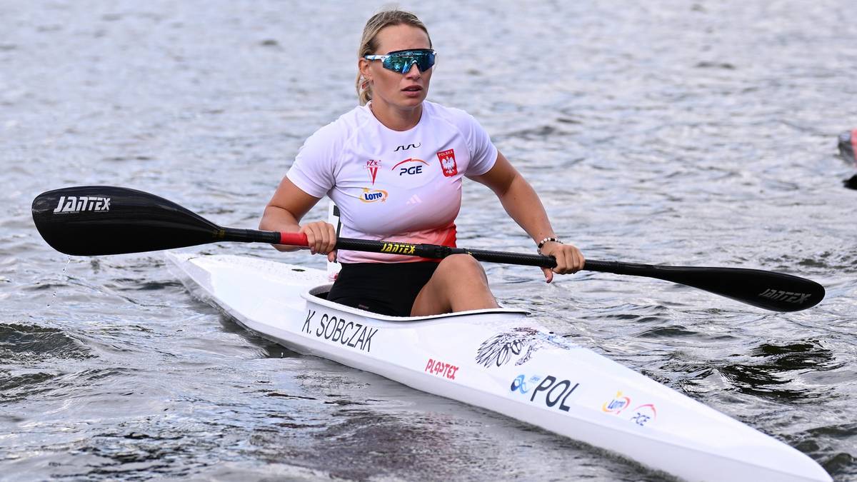 Kobieta w białej koszulce z numerem startowym i okularach przeciwsłonecznych wiosłuje w białym kajaku po wodzie.