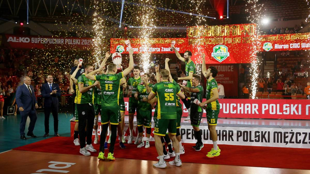 Drużyna siatkarzy w zielonych strojach świętuje zwycięstwo na tle fajerwerków, unosząc puchary.