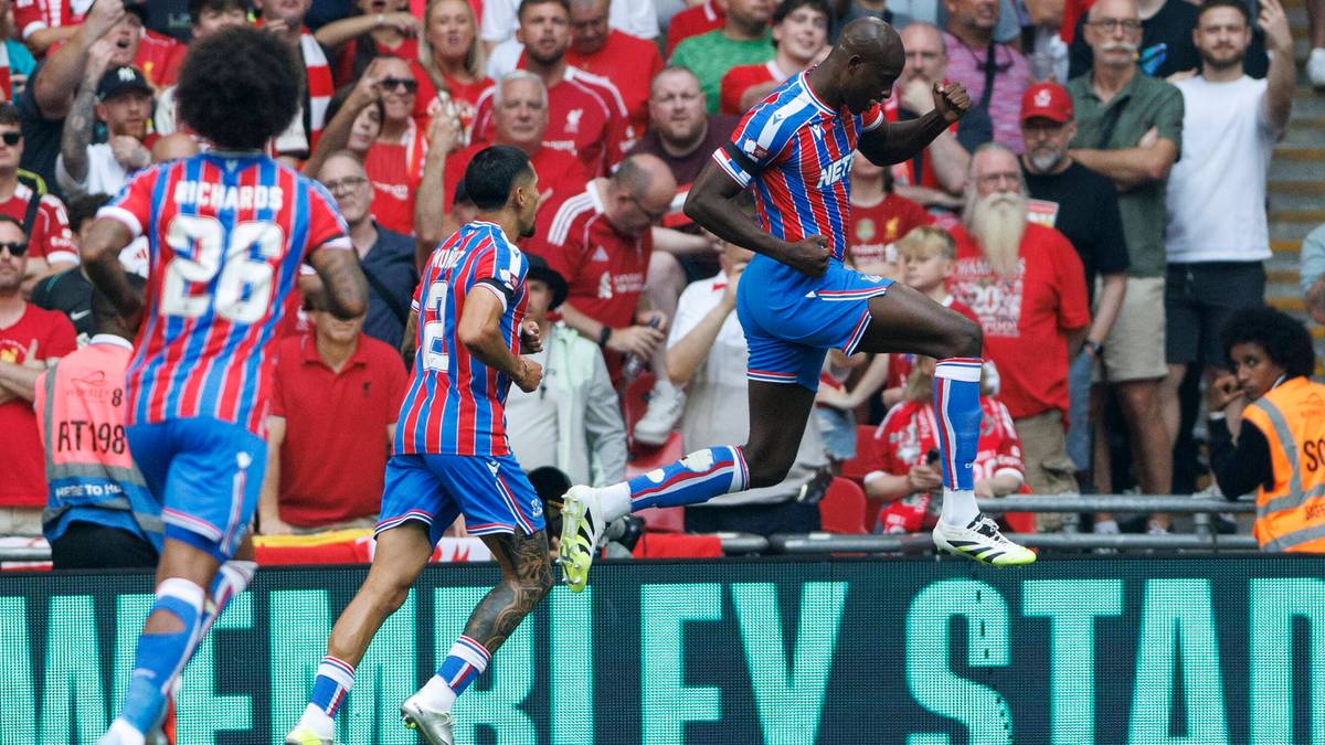 Piłkarze w czerwono-niebieskich strojach, jeden z nich zdobywa bramkę, cieszą się z drużyną i kibicami na trybunach.