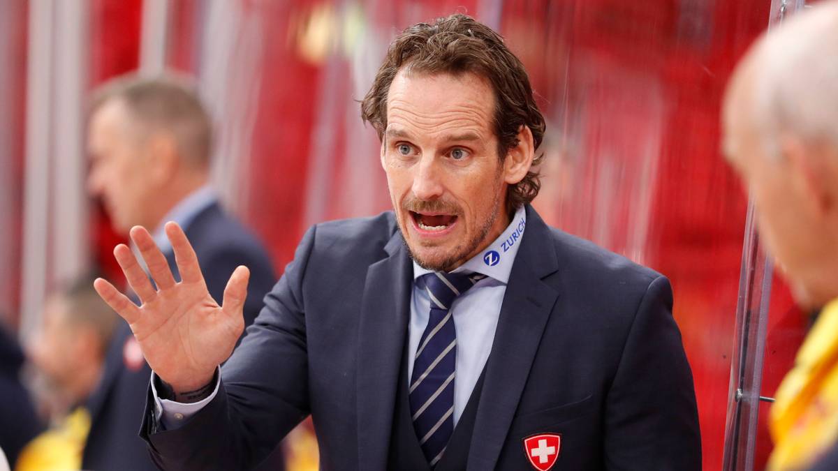Patrick Fischer, trener reprezentacji Szwajcarii w hokeju na lodzie, przemawia do zawodników w garniturze z przypinką z flagą Szwajcarii.