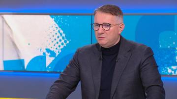 Mężczyzna w okularach i ciemnym garniturze siedzi przed ekranem z abstrakcyjnym wzorem w kolorach niebieskim i białym.