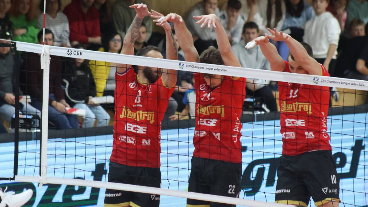 Trzech siatkarzy w czerwonych koszulkach blokuje piłkę przy siatce.