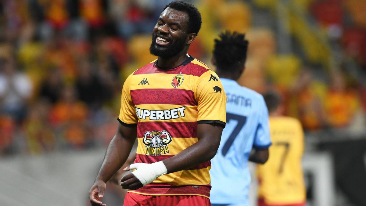 Piłkarz Jagiellonii Białystok w żółto-czerwonej koszulce uśmiecha się na boisku.