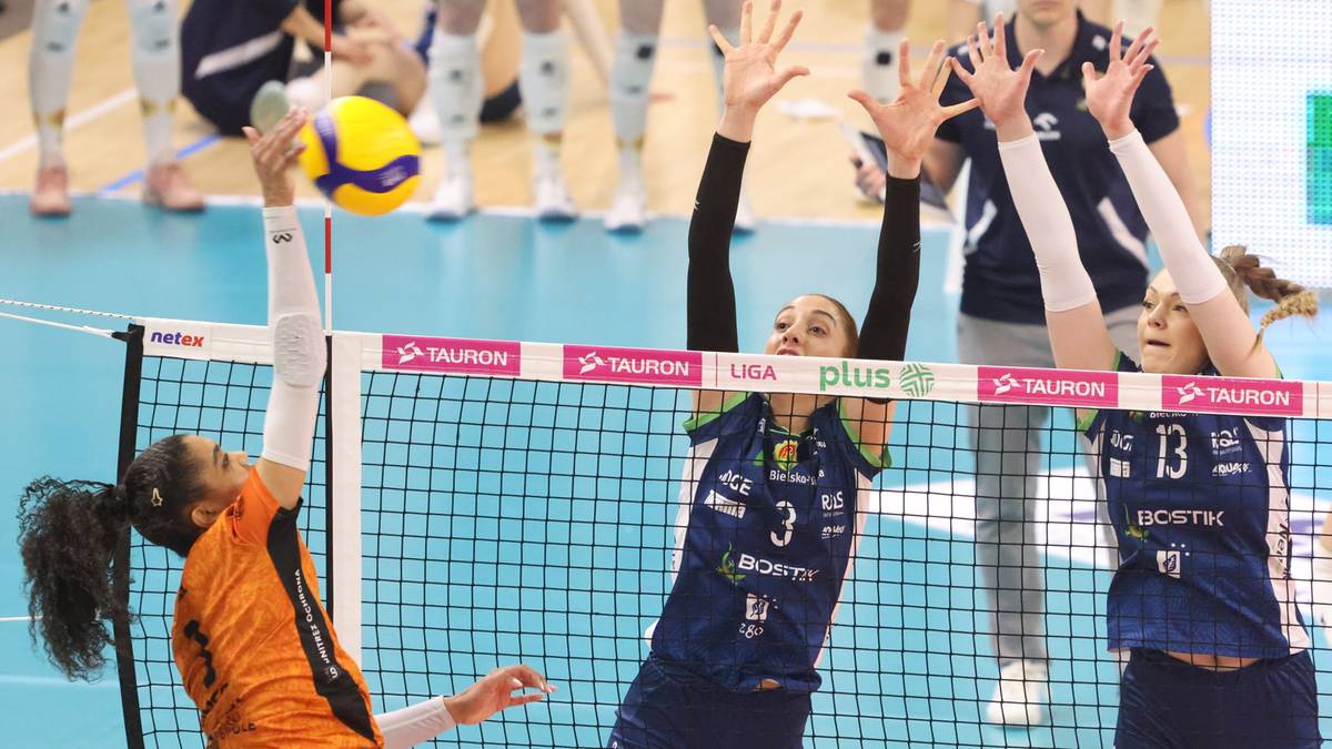 Siatkarka w pomarańczowym stroju atakuje piłkę nad siatką, podczas gdy dwie zawodniczki w niebieskich strojach pr&oacute;bują ją zablokować.