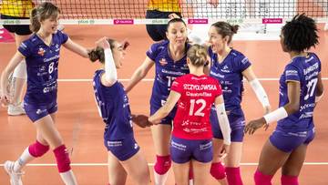 Grupa siatkarek w niebiesko-fioletowych strojach sportowych świętuje lub dyskutuje na boisku siatkarskim. Jedna z zawodniczek w czerwonym stroju z numerem 12 jest w centrum.