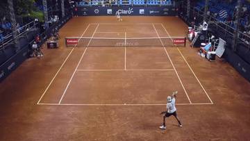 Gracz tenisa wykonuje serwis na korcie ziemnym podczas meczu. Widoczny jest drugi gracz na przeciwległej stronie kortu oraz widownia w tle.