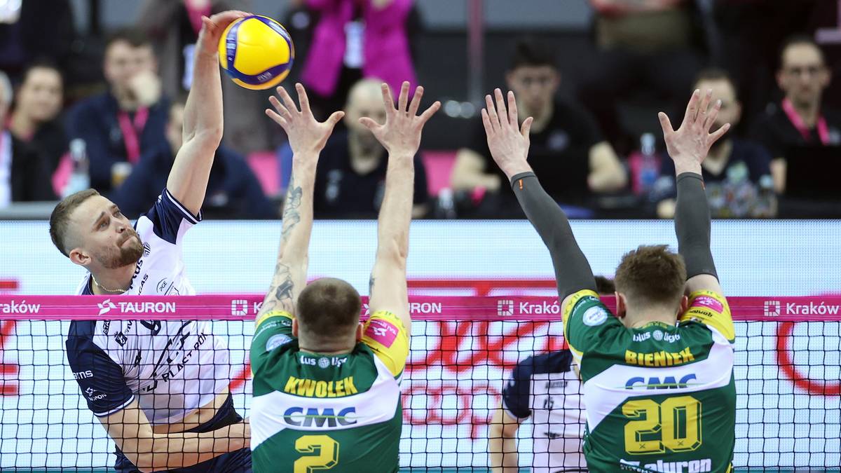 Siatkarz w białej koszulce z niebieskimi elementami, w akcji stara się odbić piłkę, blokują go dwaj siatkarze w zielonych koszulkach z żółtymi elementami.