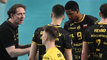 Trener wpatruje się w zawodników w czarnych strojach, gestykulując i udzielając im wskazówek podczas przerwy w meczu siatkówki.