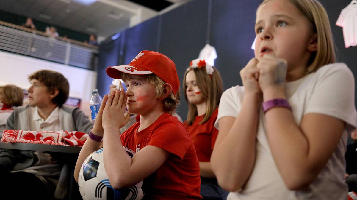 Grupka dzieci z flagami i malowanymi twarzami na twarzach, w skupieniu ogląda mecz piłki nożnej.
