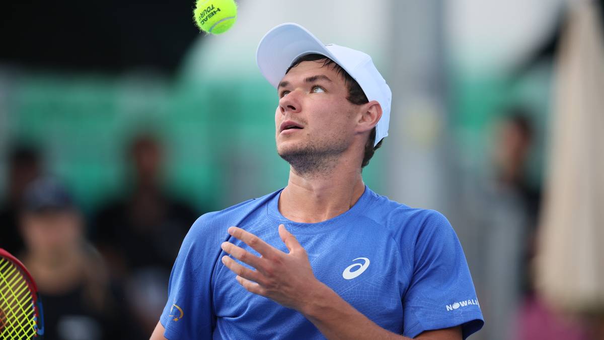 Tenisista w niebieskiej koszulce i białej czapce patrzy w górę na lecącą piłkę tenisową.