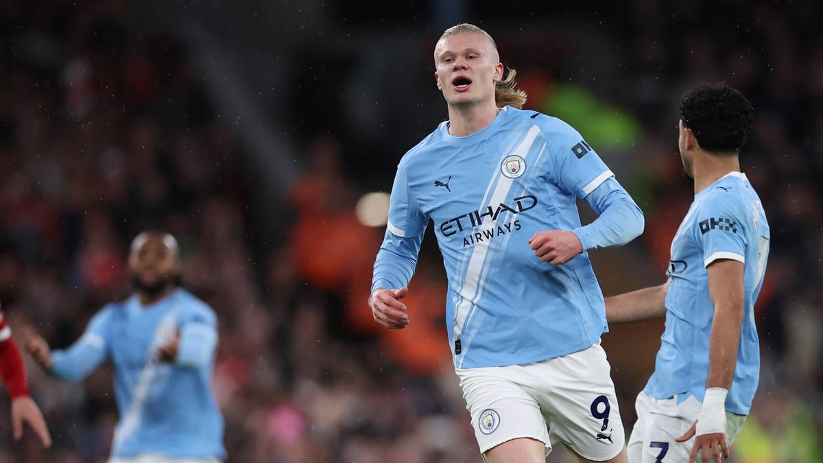 Piłkarz Manchesteru City Erling Haaland z podniesioną ręką, w tle inny piłkarz w jasnobłękitnej koszulce.