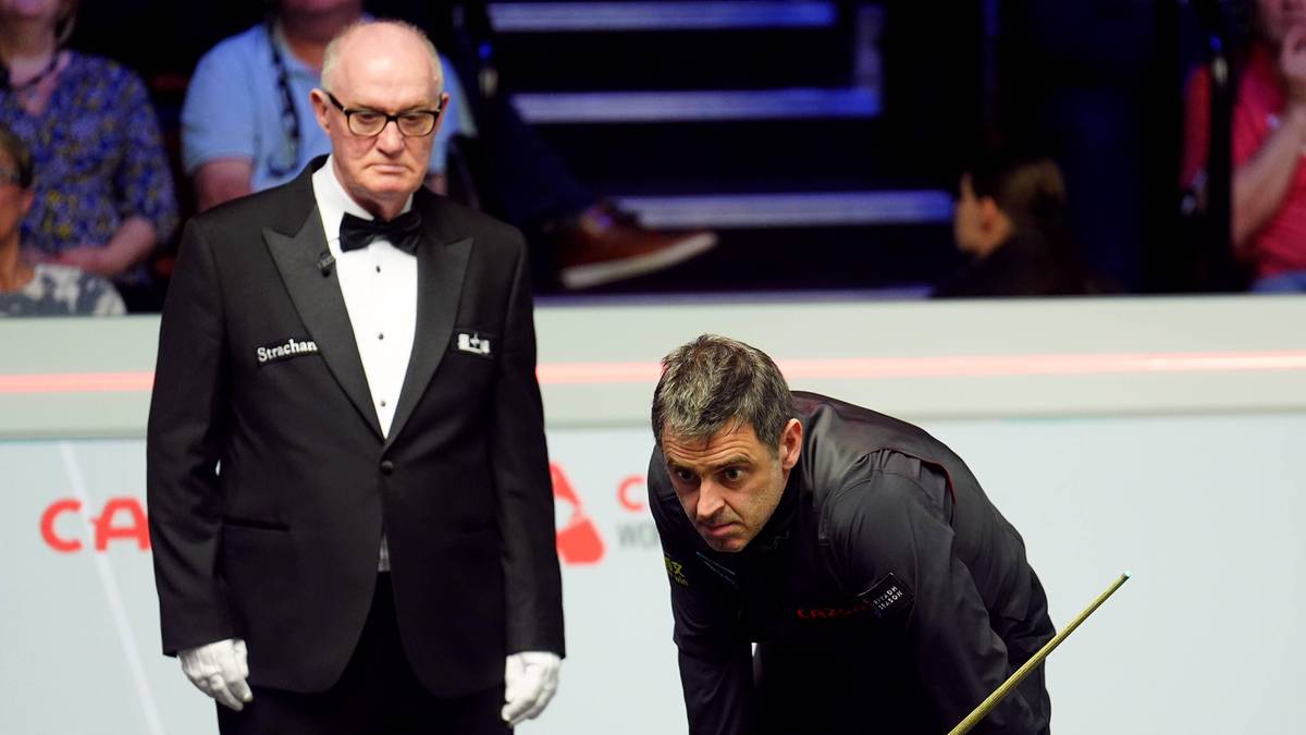 Sędzia snookerowy w czarnym fraku i białych rękawiczkach stoi obok zawodnika w czarnej koszulce, kt&oacute;ry przygotowuje się do uderzenia kijem w bilę na zielonym stole.