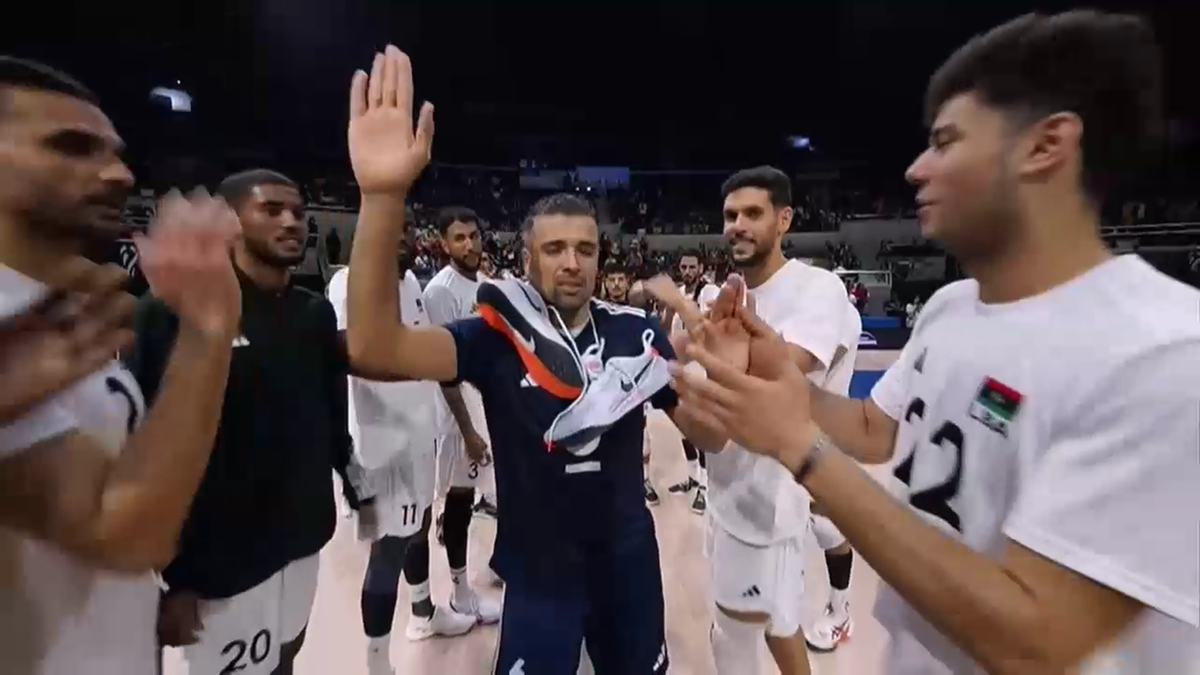 Grupa koszykarzy w strojach meczowych gratuluje kapitanowi, który trzyma but w ręce i podnosi drugą dłoń.